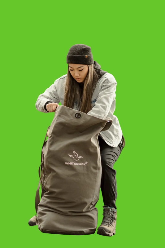 Person sitting on a log in a forest, opening a large bag with 'White Duck' branding.