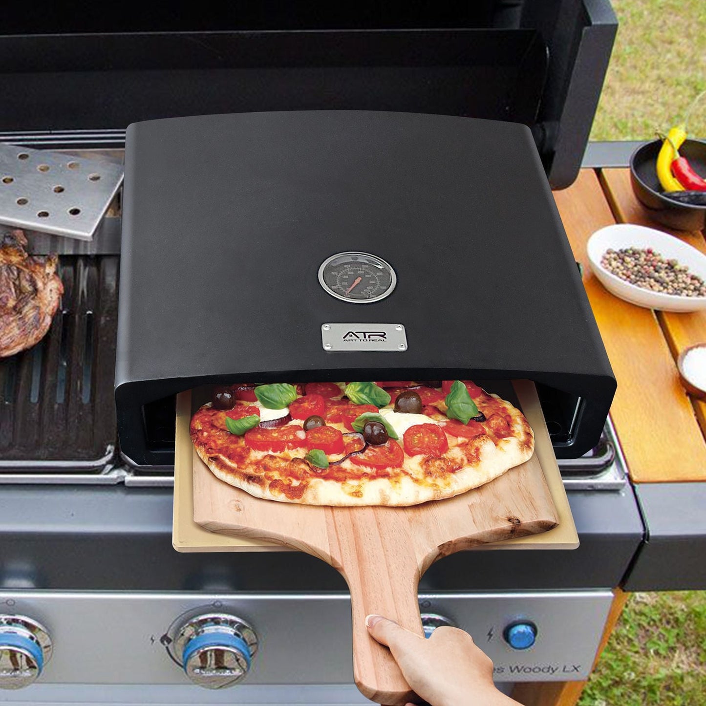 Person placing a pizza into a portable pizza oven on a grill outdoors.