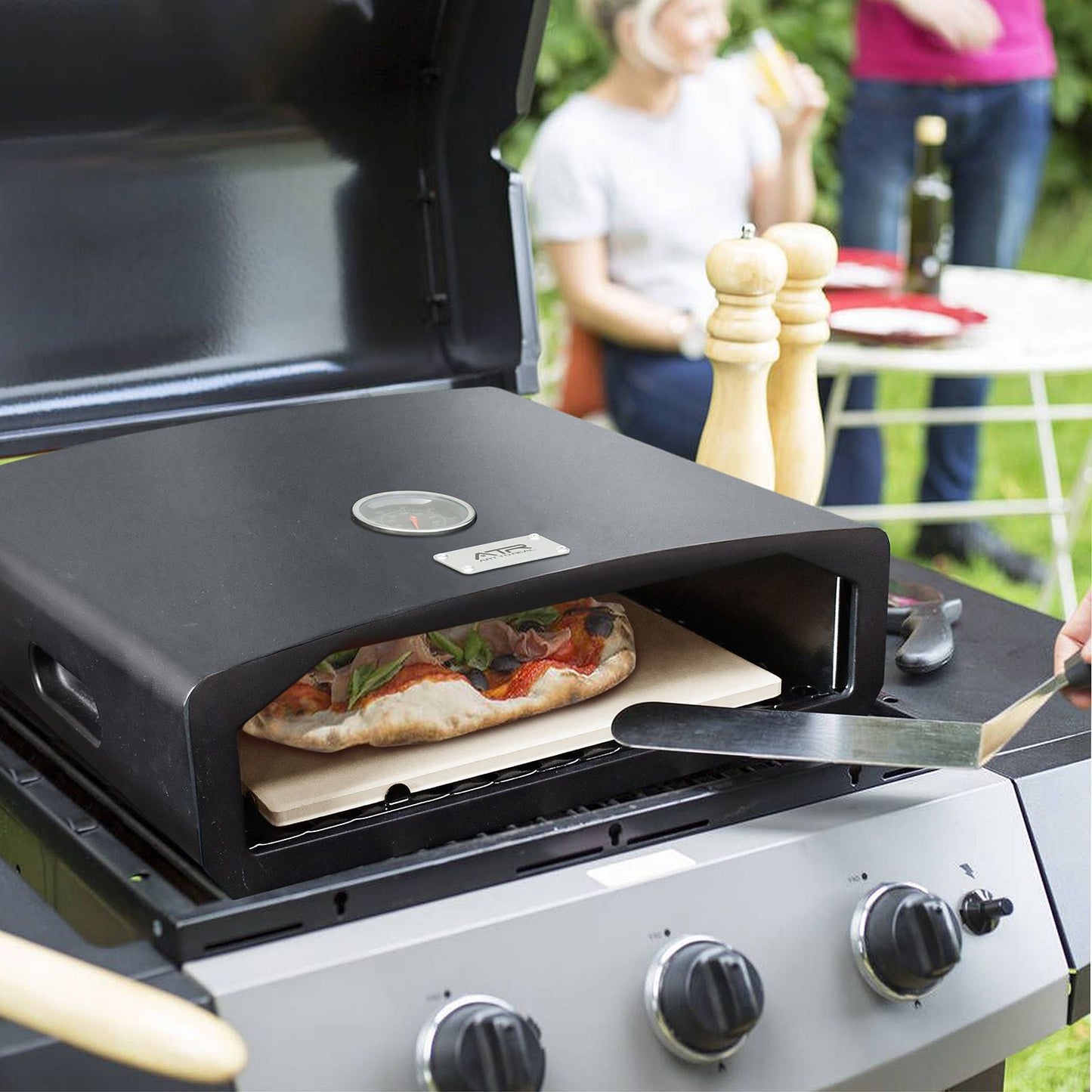 Portable pizza oven attached to a grill with a pizza being cooked, people in the background.