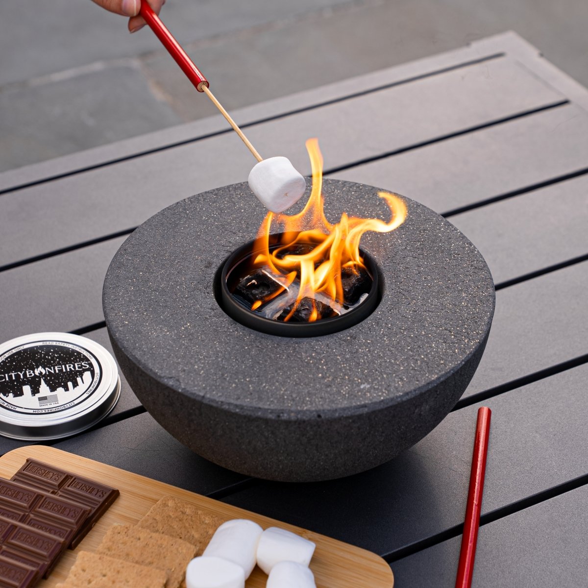 Person roasting marshmallows over a portable fire pit on a wooden surface.