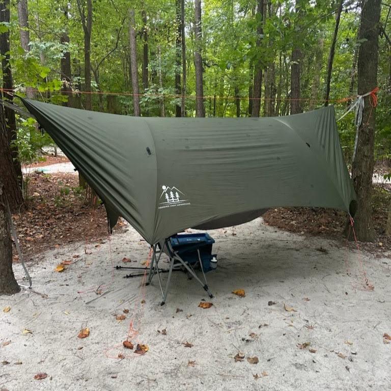 Green camping tarp set up in a forested area with trees and ground cover visible.