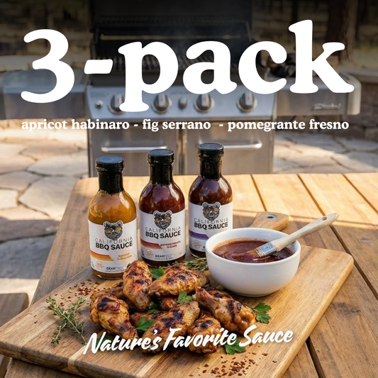 Three bottles of barbecue sauce with a bowl of sauce and grilled chicken on a wooden table outdoors.