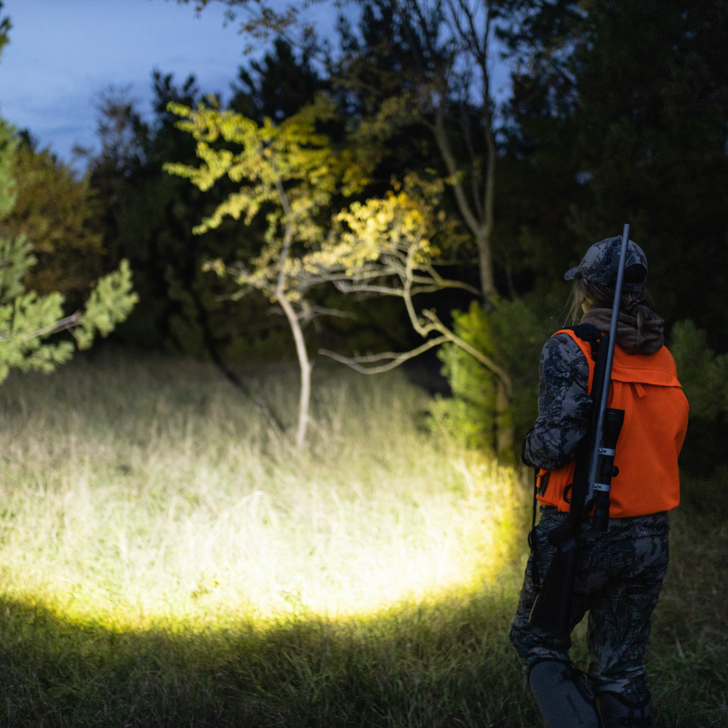 Person in camouflage clothing and orange vest holding a flashlight in a dark forest setting