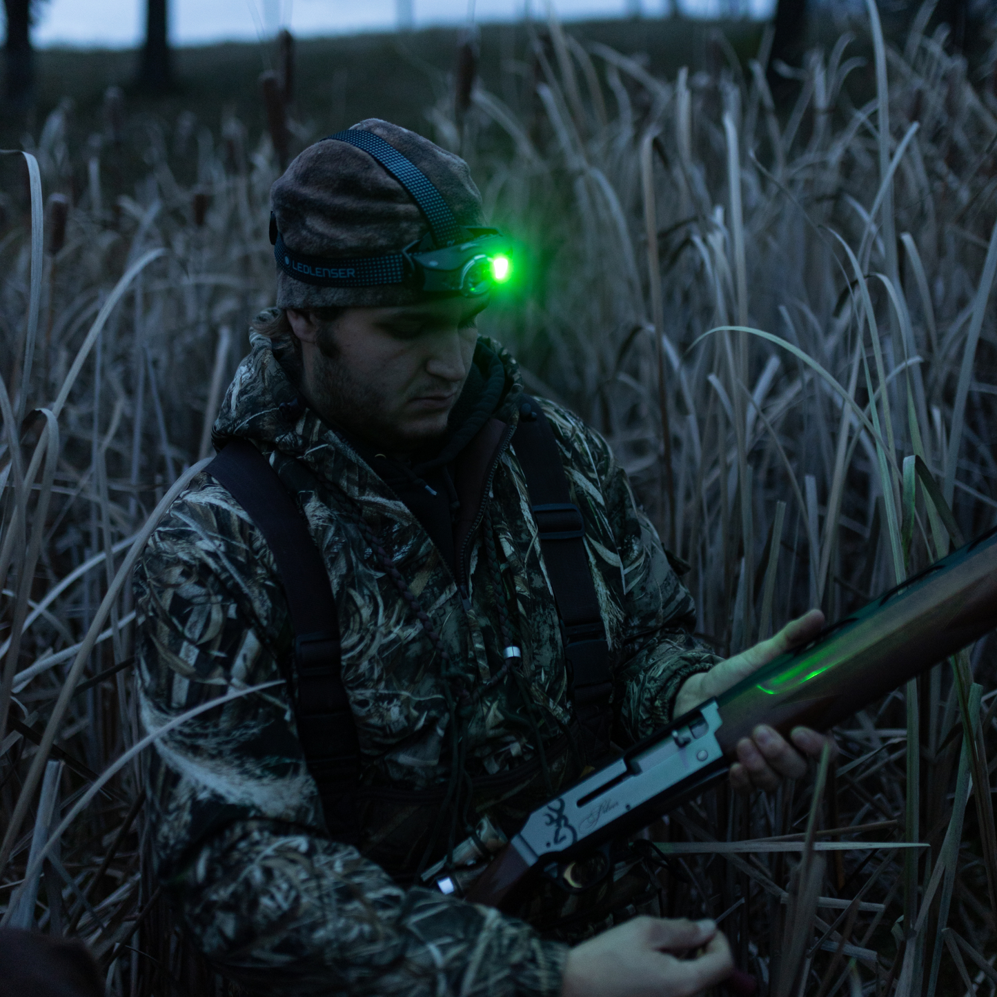 Hunter in camouflage gear with a rifle and headlamp in tall grass at night.