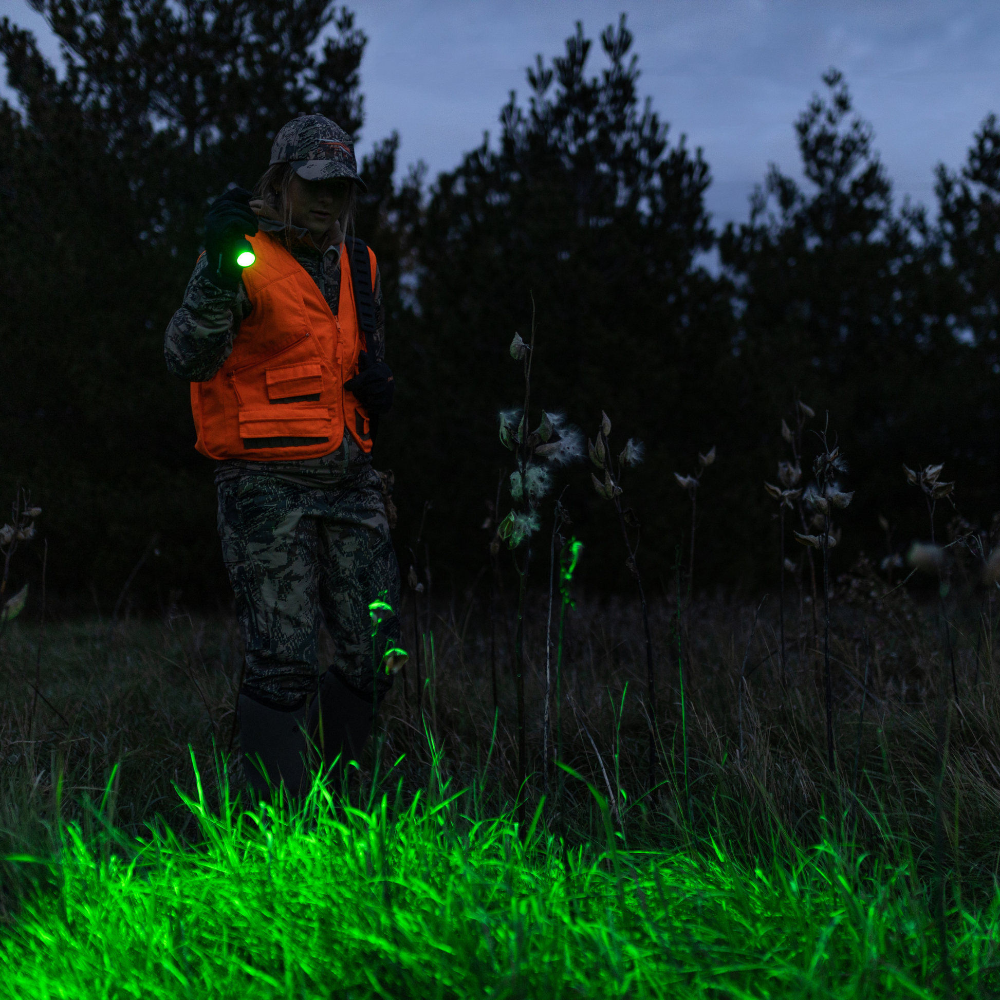 Person in orange vest and camouflage pants holding a green light source in a dark forest setting.