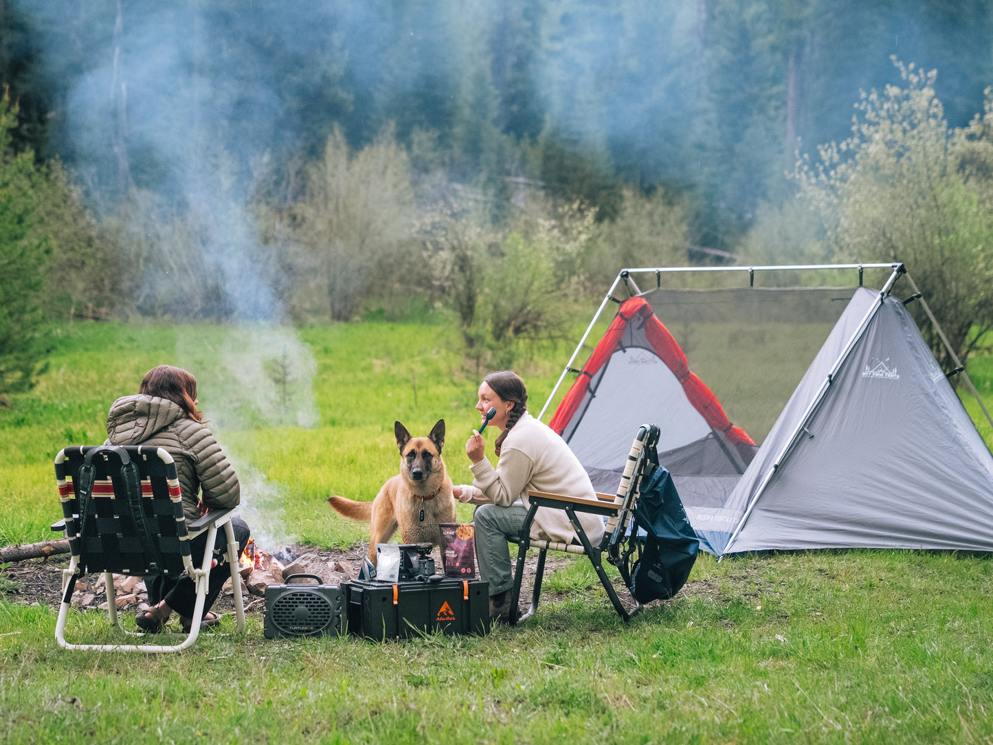 Two people and a dog sitting by a campfire near a tent in a forest setting.