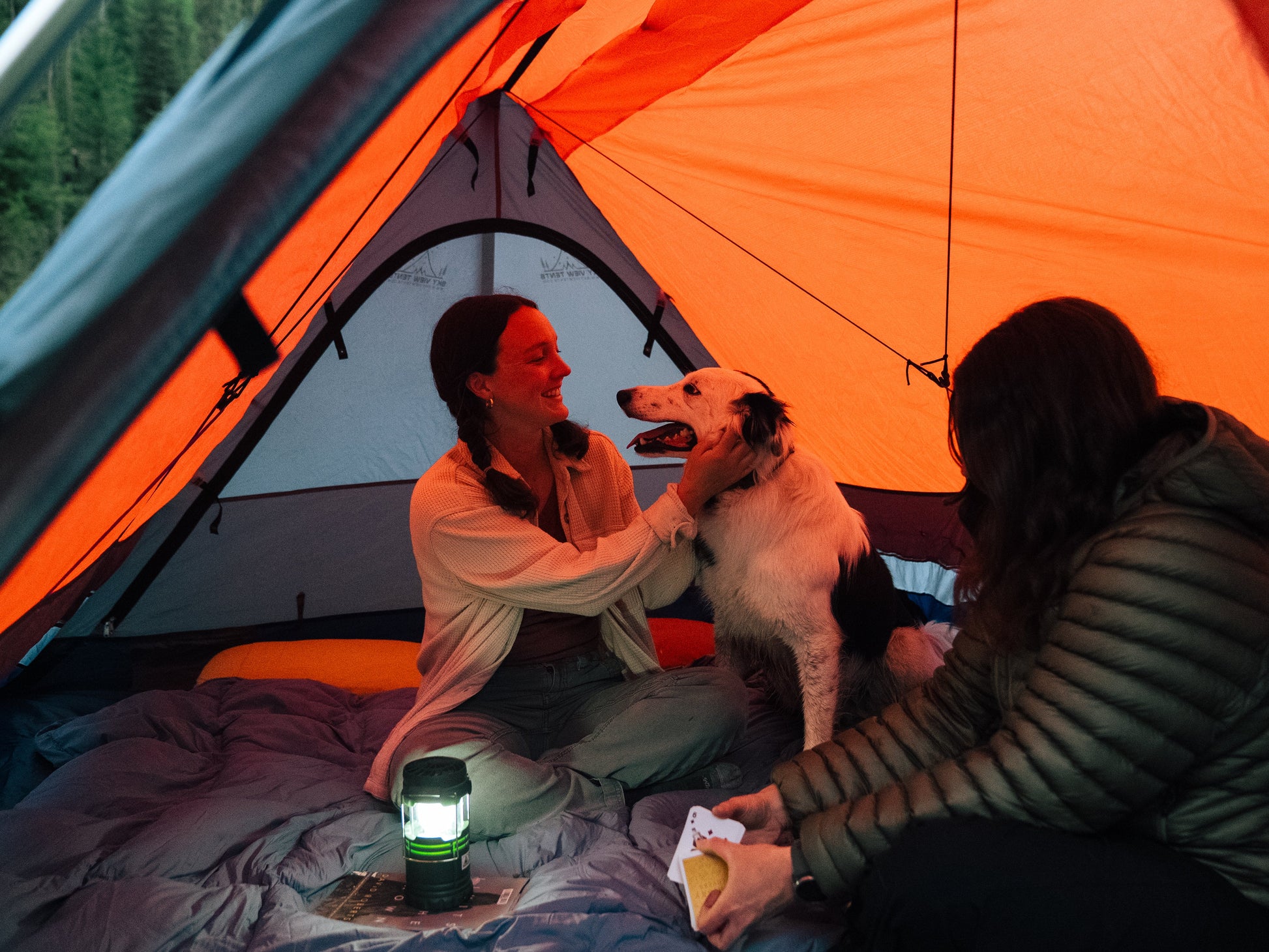 Two people inside an orange tent with a dog, holding a snack.