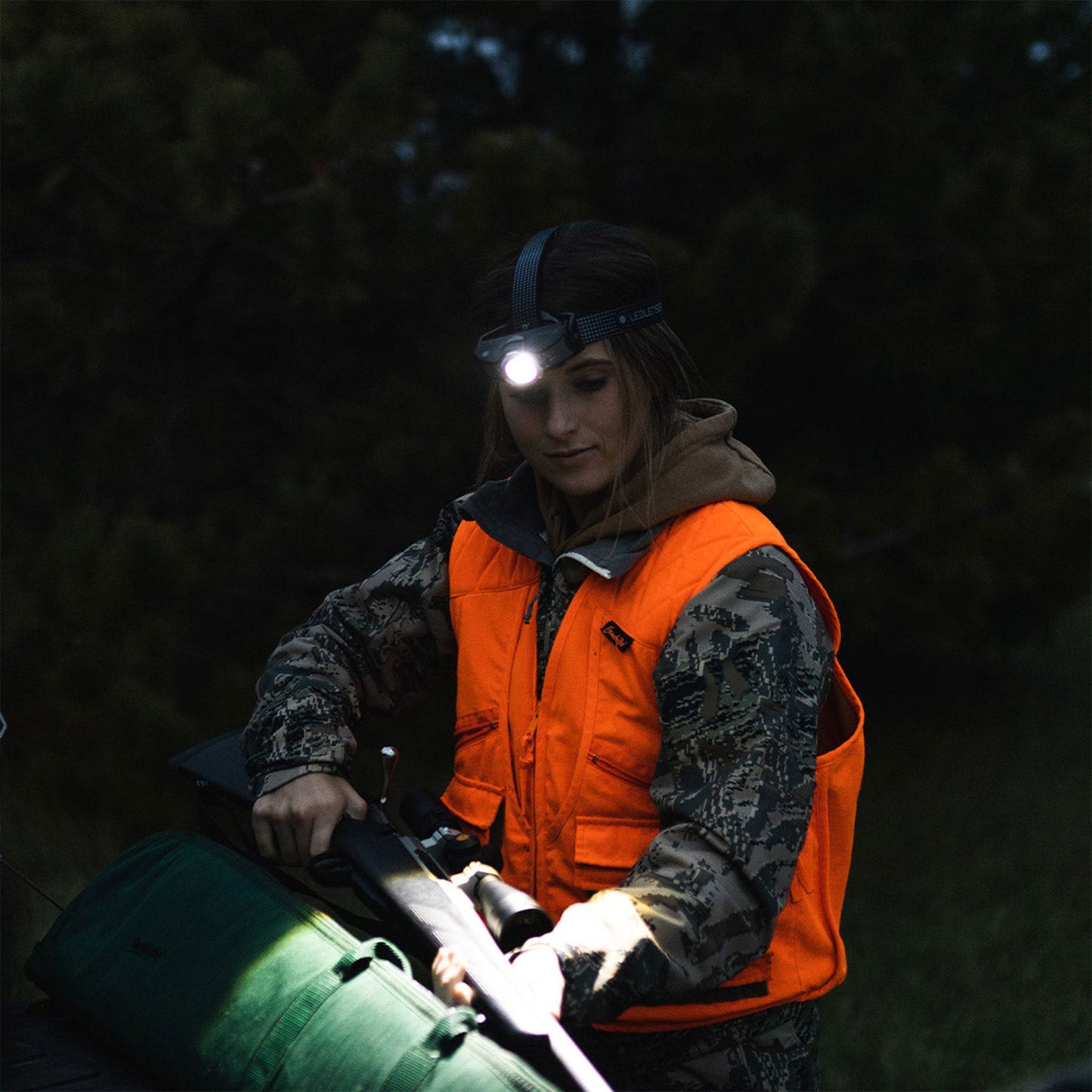 Person in camouflage jacket and orange vest with headlamp working on a green object in a dark forest.