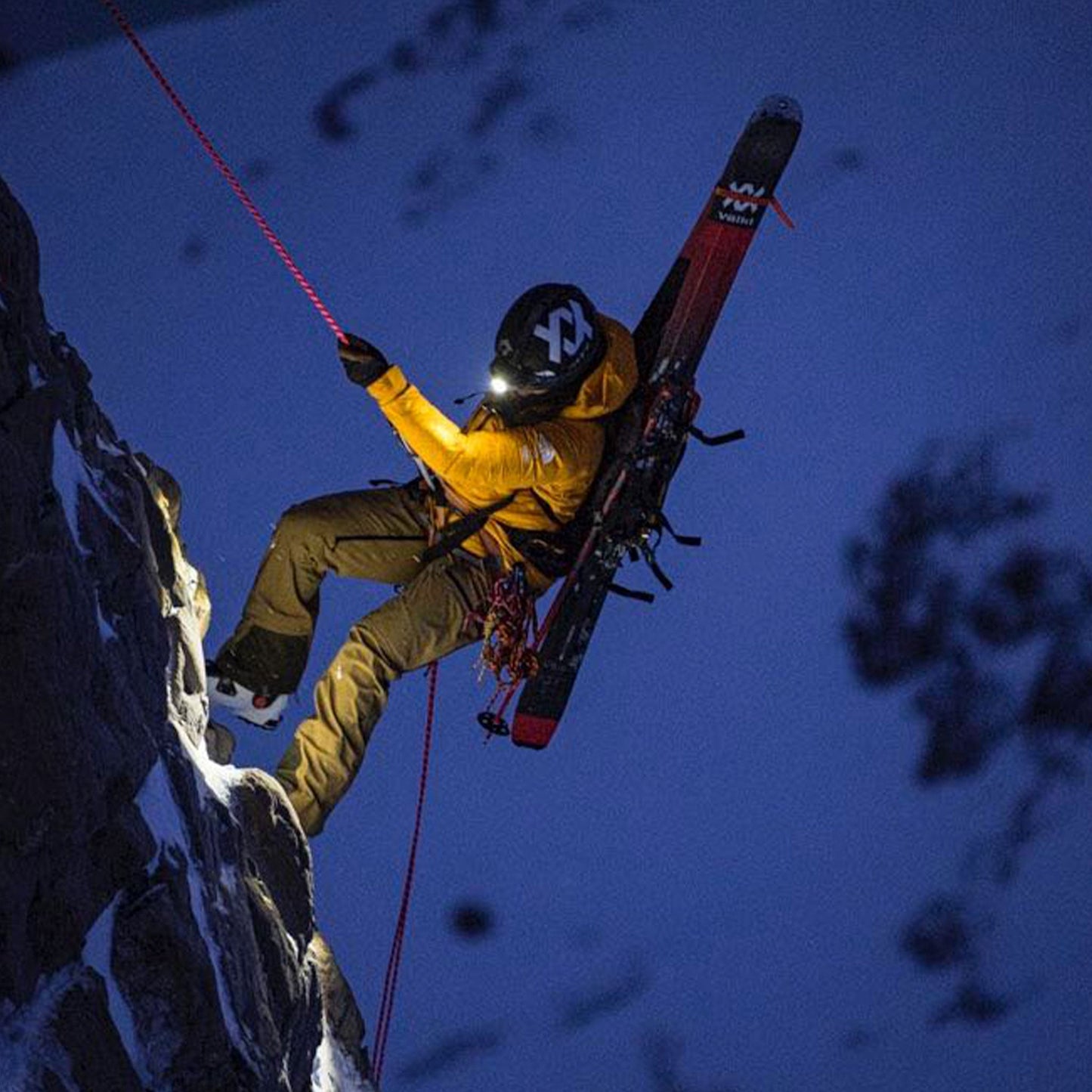 Person in yellow jacket and helmet with skis, climbing a snowy mountain at night.