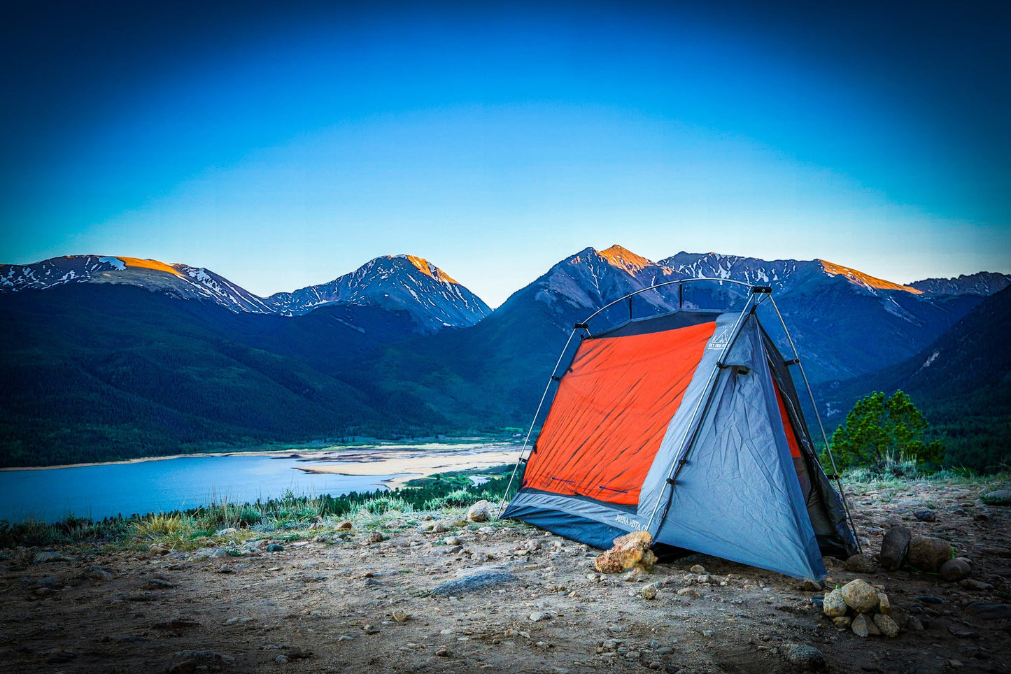 A red and gray camping tent pitched on a rocky clearing in front of a mountain landscape with a lake and blue sky.