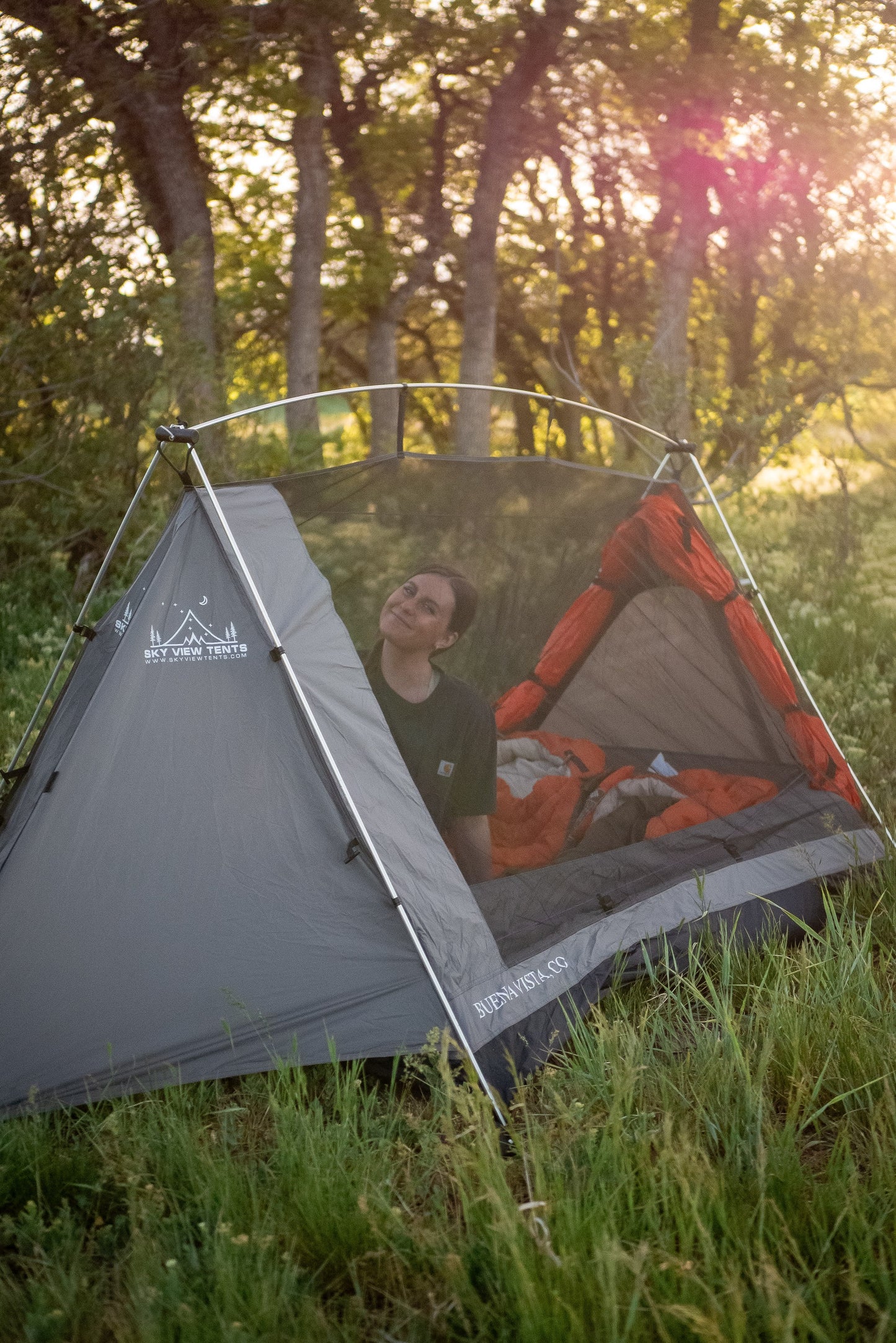 A gray and orange camping tent with a mesh door is pitched in a grassy forest clearing at sunset, with a person sitting inside looking upward.