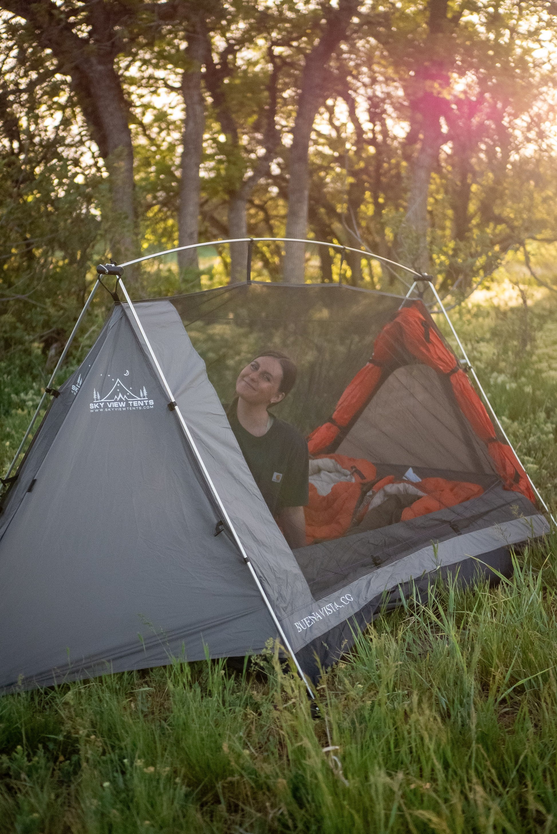 A gray and orange camping tent with a mesh door is pitched in a grassy forest clearing at sunset, with a person sitting inside looking upward.