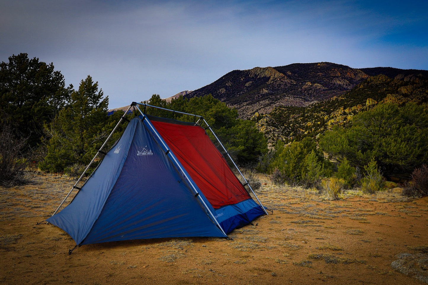 Blue and red tent set up in a desert landscape with mountains and trees in the background.
