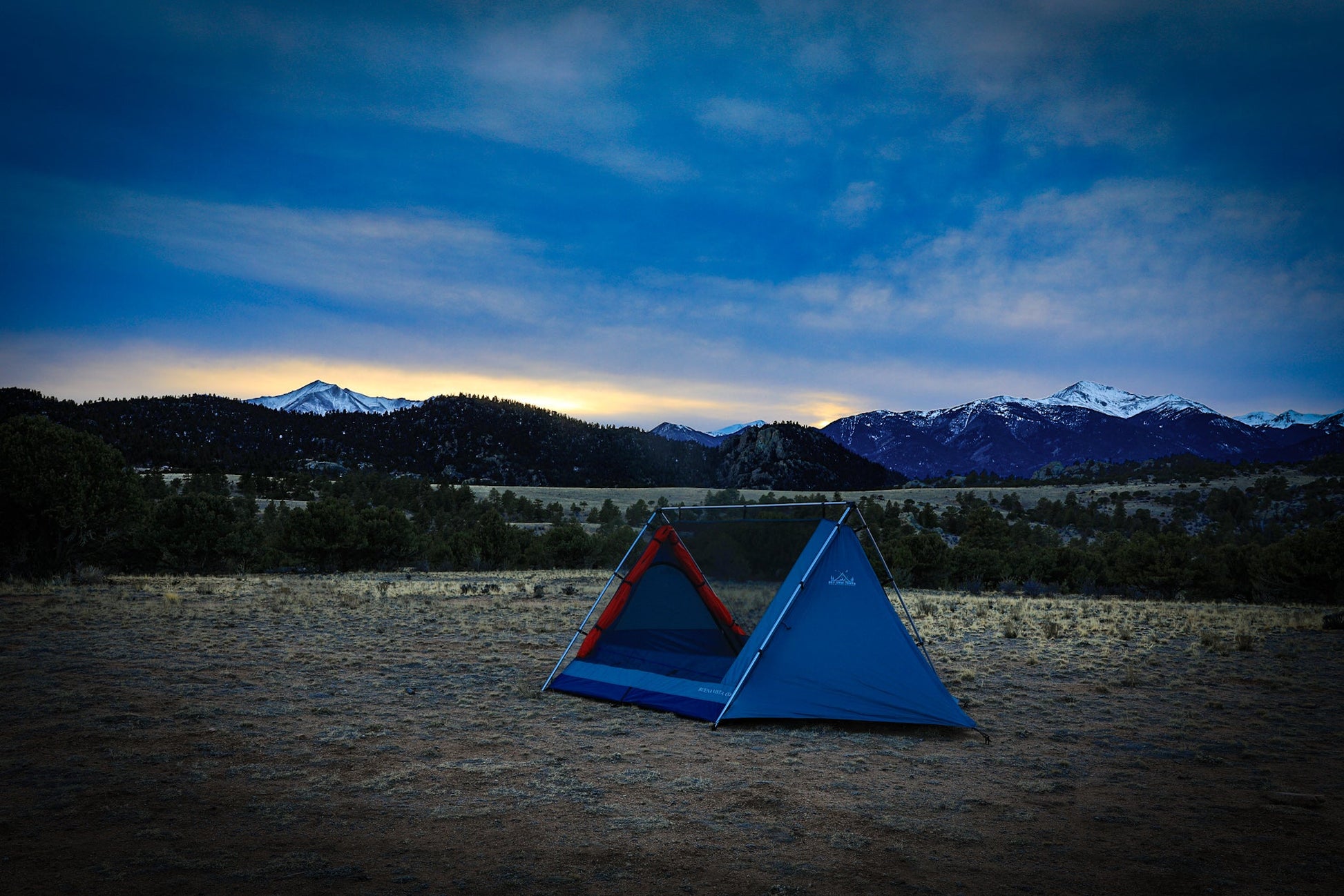 Two triangular tents on a sandy ground with mountains in the background during sunset.