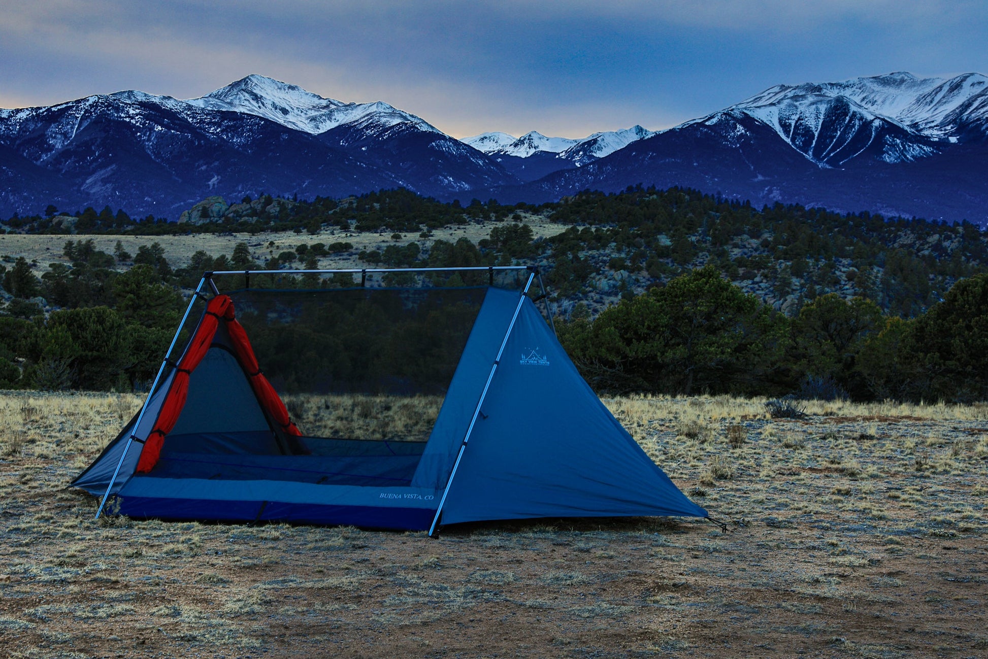 Blue and red tent set up in a field with snow-capped mountains in the background