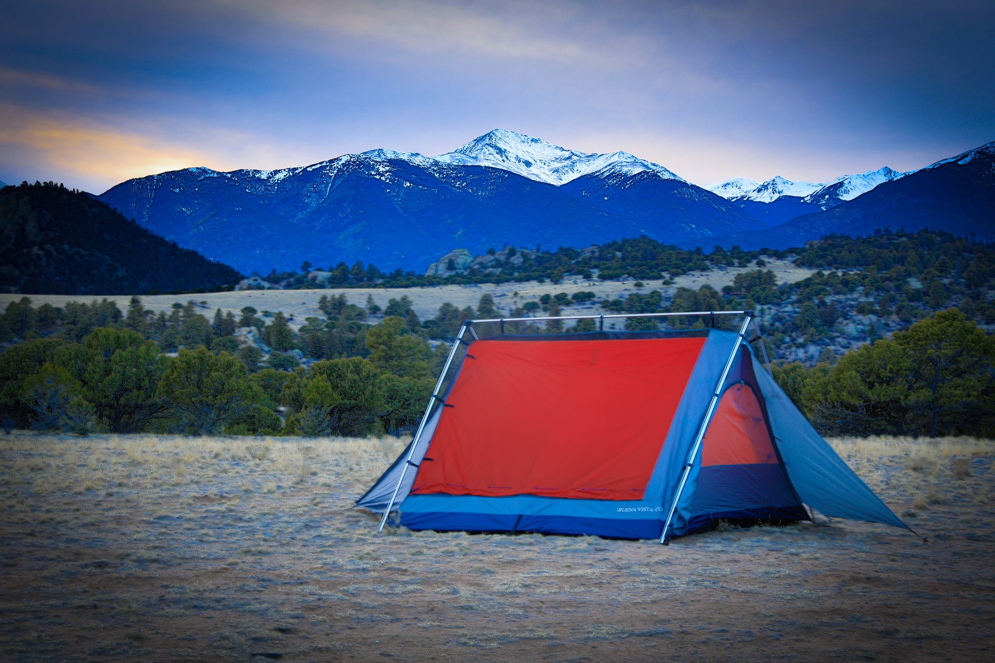 Red and blue tent set up in a field with mountains in the background