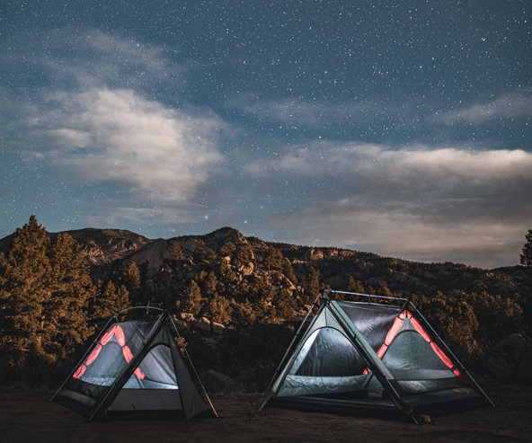 Two camping tents pitched on a mountain clearing under a starry night sky with clouds and dark rocky peaks in the background.
