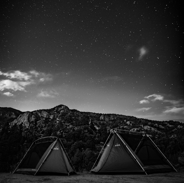 Two dark camping tents pitched in front of rocky mountains under a star-filled night sky in black and white.