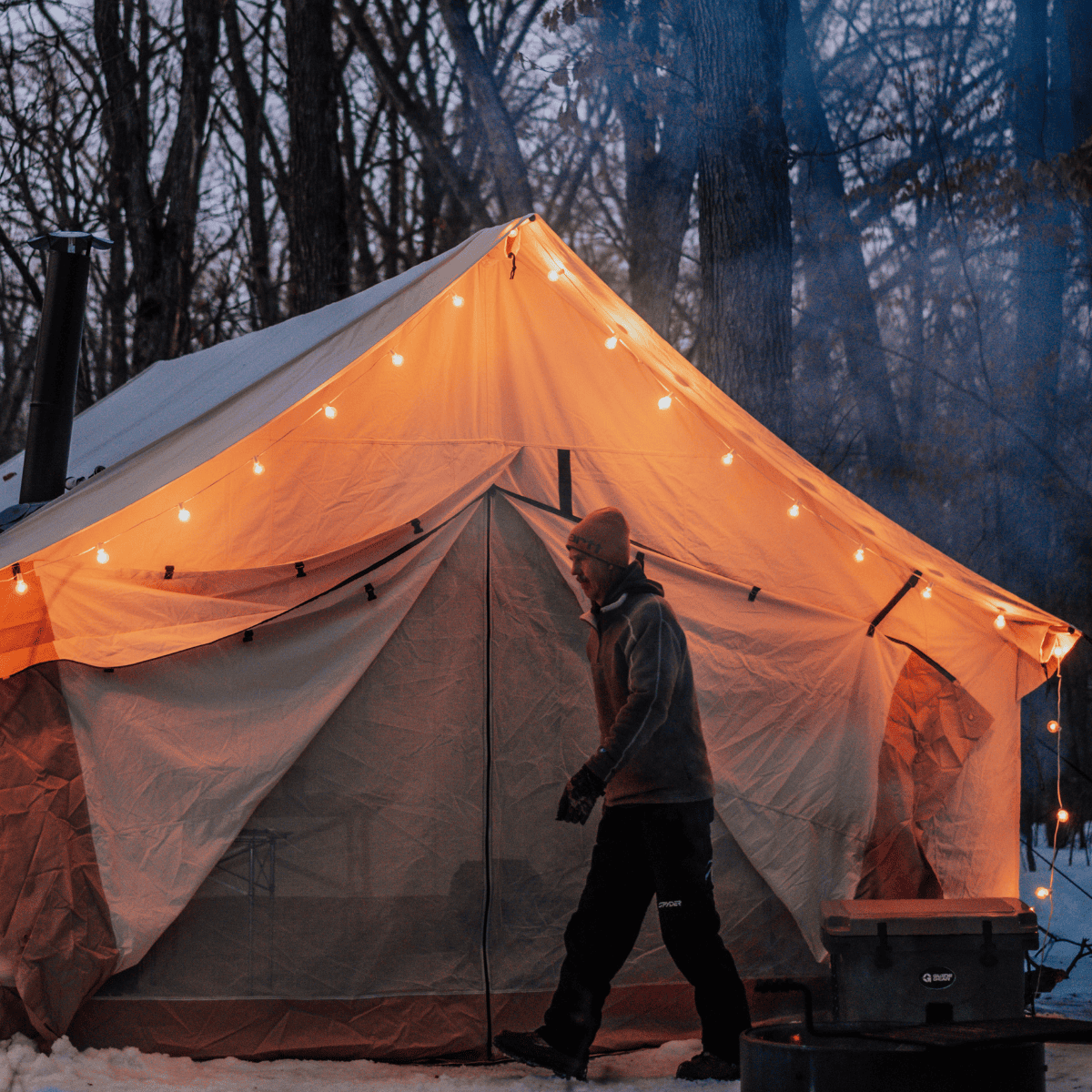 Person walking towards a tent with string lights in a snowy forest