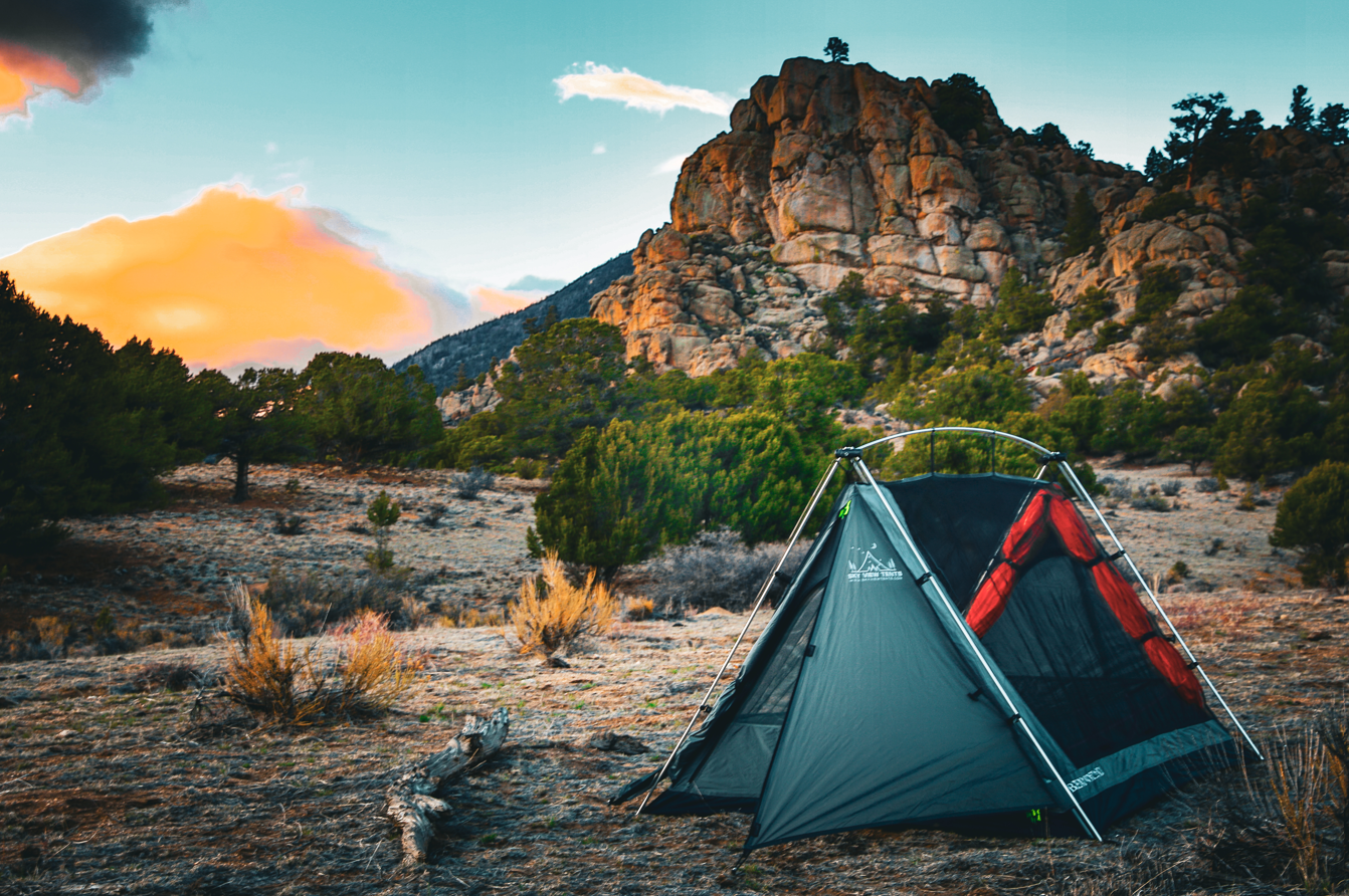 A dark green and orange tent pitched in a rocky desert landscape at sunset, with trees, shrubs, and a large rock formation in the background.