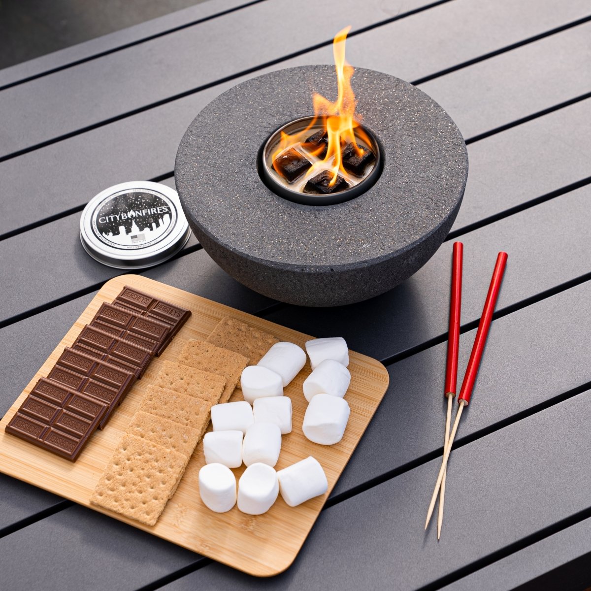 Fire pit table with flames, chocolate bars, graham crackers, marshmallows, and sticks on a wooden surface.