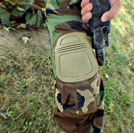 Person wearing camouflage pants with a knee pad holding a gun outdoors.