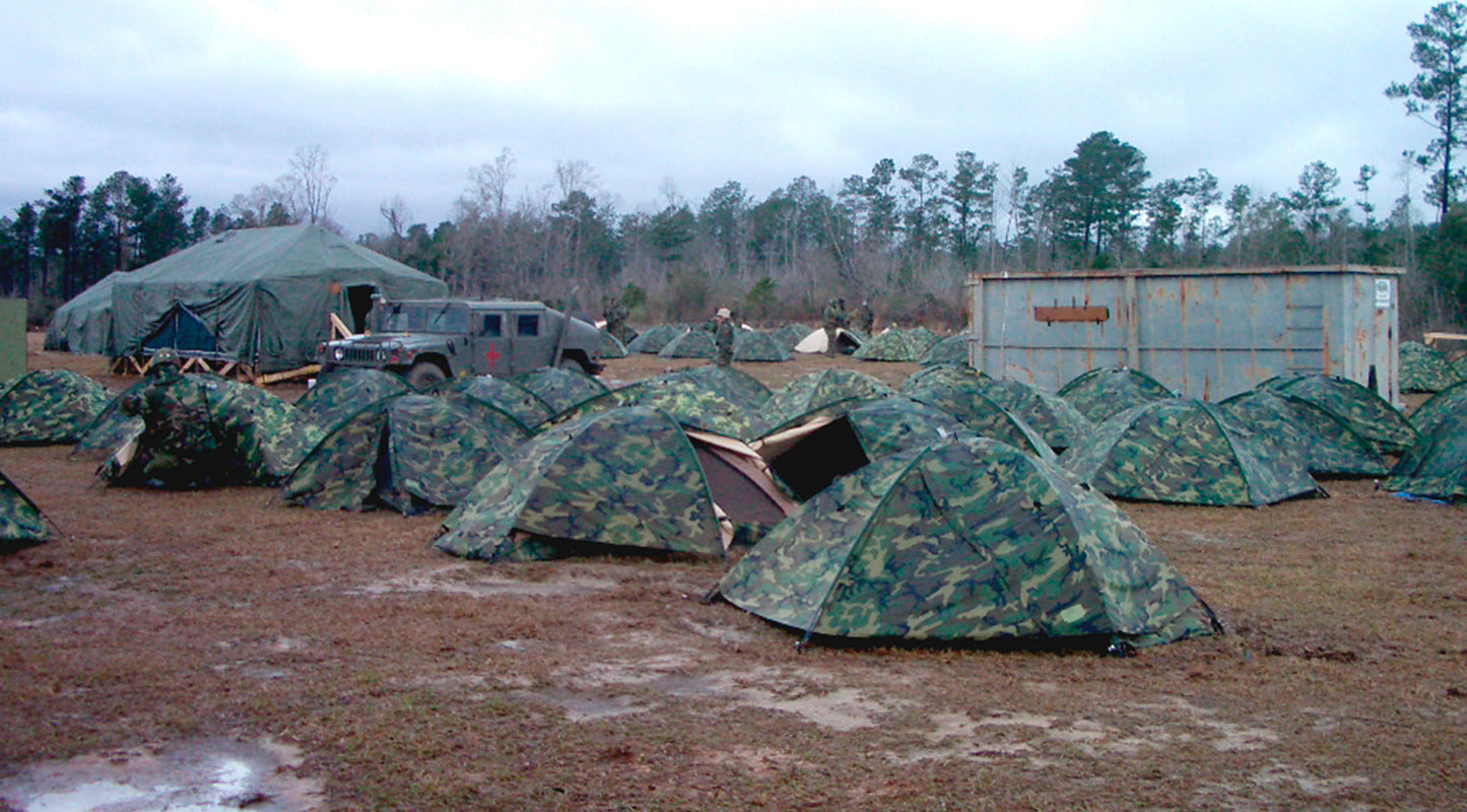 Camouflage tents set up in a field with a truck and storage containers in the background.