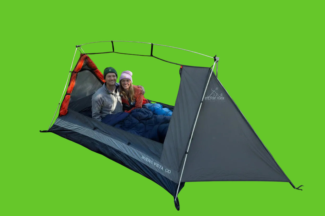 A couple relaxes inside a gray and orange Kenda Forest Co. dome tent set up on a dirt clearing in a forest.