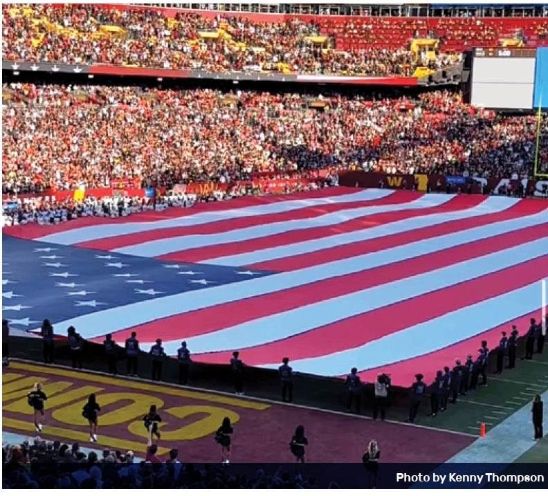 Large American flag displayed on a sports field with spectators in the stands.